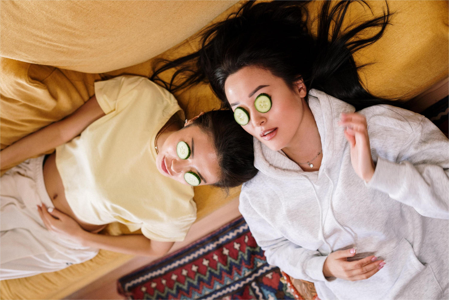 two women with cucumber's on their eyes laying on sofa